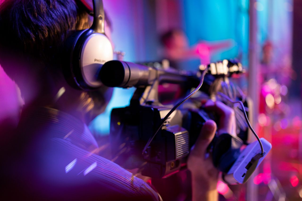 A videographer with headphones focuses intently while filming an indoor event under colorful lights.